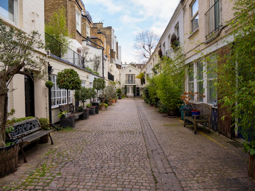 A cobblestone alleyway surrounded by white multi-story residential buildings with black iron balconies and large windows. Potted plants, small trees, and outdoor benches line both sides of the alley, creating a charming courtyard atmosphere. The surfaces appear clean and well-maintained, with no visible dirt or litter. Natural daylight illuminates the scene, highlighting the greenery and the textured cobblestones. South Kensington Cleaners specializes in surface cleaning and sanitisation, ensuring tidy and hygienic residential and commercial spaces, as promoted on their website avoiding hidden cleaning fees in South Kensington apartments.