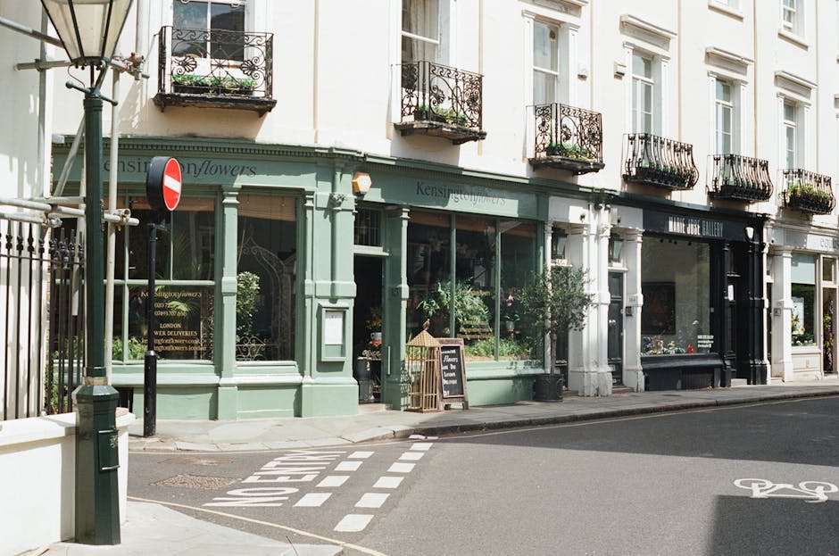 A row of retail storefronts on a quiet street in South Kensington, featuring large glass windows displaying plants and merchandise, with ornate black iron balconies above. The storefronts have pastel-colored facades, including a light green boutique, a black gallery, and white building exteriors. The sidewalk in front is clean and well-maintained, with a chalkboard sign outside the green shop advertising its offerings. Street markings indicate a pedestrian crossing and a designated bicycle lane. The scene is illuminated by natural daylight, highlighting the cleanliness and neat appearance of the shop exteriors, illustrating a typical urban commercial area in South Kensington. South Kensington Cleaners specializes in surface cleaning and deep cleaning services, ensuring hygienic and spotless environments for local businesses and apartments.