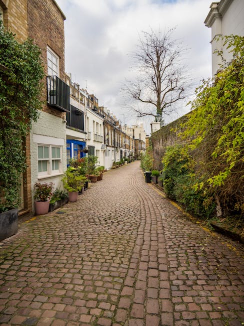 A cobblestone alleyway surrounded by white multi-story residential buildings with black iron balconies and large windows. Potted plants, small trees, and outdoor benches line both sides of the alley, creating a charming courtyard atmosphere. The surfaces appear clean and well-maintained, with no visible dirt or litter. Natural daylight illuminates the scene, highlighting the greenery and the textured cobblestones. South Kensington Cleaners specializes in surface cleaning and sanitisation, ensuring tidy and hygienic residential and commercial spaces, as promoted on their website avoiding hidden cleaning fees in South Kensington apartments.