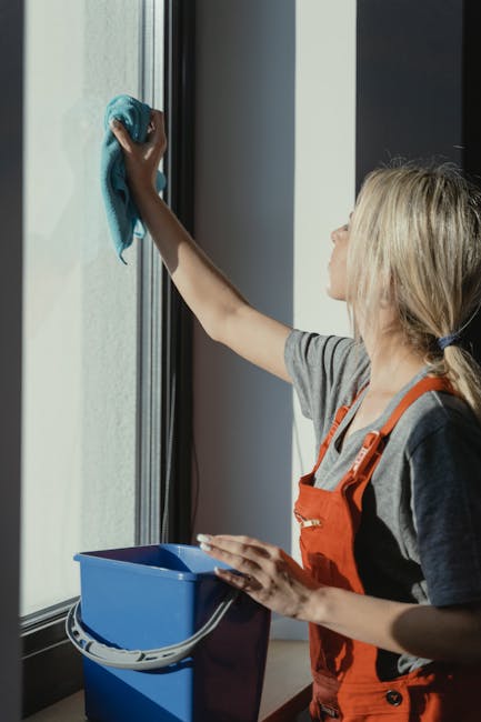 A woman with blonde hair tied back, wearing a grey shirt and an orange overall, is cleaning a large glass window with a blue cloth. She is holding a blue spray bottle in her left hand, with a white microfiber cloth in her right hand, which she is using to wipe the window surface. The window frame is black, and natural light is illuminating the scene, highlighting the clean and clear glass. The woman appears focused on the task, with a blue bucket placed on the floor nearby. This setting illustrates surface cleaning and window sanitation process, reflecting domestic cleaning methods in a residential environment, consistent with services offered by South Kensington Cleaners, as discussed on the page about weekend cleaning in SW7, South Kensington.