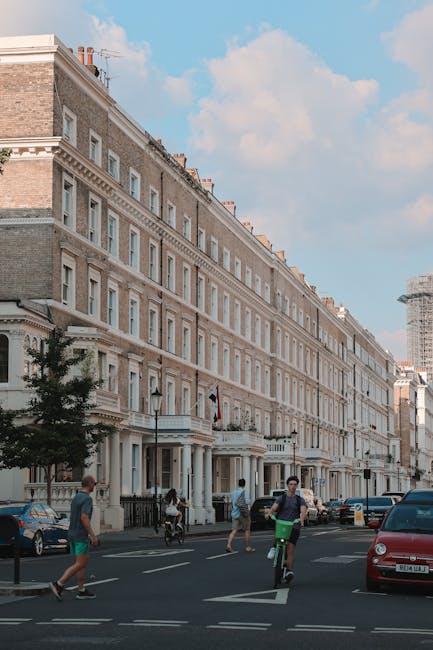 Photograph of a street scene outside a historic, multi-storey Victorian building in South Kensington, with elegant stone facades, large sash windows, and decorative architectural details. The street features parked cars along the curb and pedestrians, including a person riding a bicycle, walking on the sidewalk. The scene is illuminated by natural daylight with a partly cloudy sky overhead. This image emphasizes the importance of surface cleaning and maintenance for exterior building surfaces and nearby areas, aligning with the residential and commercial cleaning services provided by South Kensington Cleaners, as highlighted in the Exhibition Road SW7 cleaning guide for South Kensington flats.