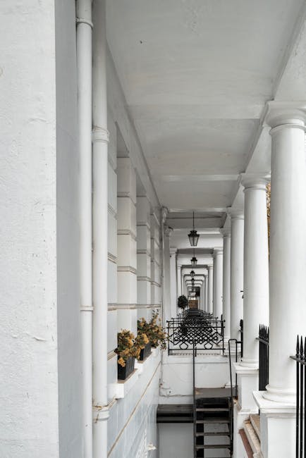 A narrow, covered outdoor corridor in a residential building with white-painted walls, columns, and a ceiling. The space features black wrought iron railings and decorative lamp fixtures hanging from the ceiling. Potted plants with flowers are positioned along the wall, contributing to an inviting atmosphere. The flooring appears to be a clean, smooth surface, possibly stone or concrete. The area is well-maintained and tidy, with no visible dust or dirt, exemplifying professional surface cleaning and maintenance typical of residential spaces. South Kensington Cleaners specializes in deep cleaning and sanitisation services, ensuring such exteriors are kept pristine and hygienic.