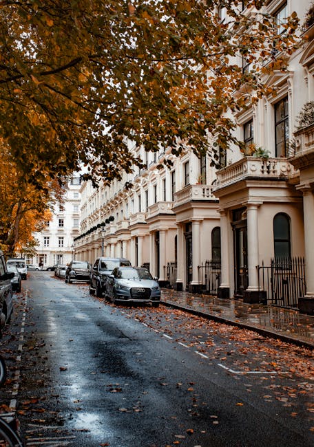 A residential street in South Kensington featuring a row of white Victorian-style terraced houses with decorative moldings and black wrought iron fences. The pavement is wet, reflecting the overcast sky, and fallen autumn leaves are scattered across the road and sidewalk. Several parked cars are along the curb, and a large tree with orange and brown leaves extends its branches over the street, creating a canopy. The scene is quiet with no pedestrians visible. The image illustrates a tranquil urban setting in need of surface cleaning and maintenance, which South Kensington Cleaners offers through comprehensive domestic cleaning and deep cleaning services in the area.
