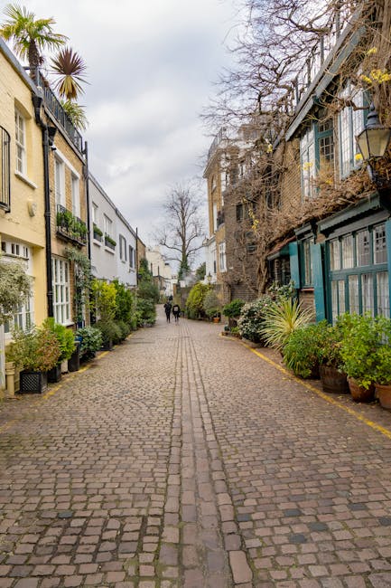 A cobblestone residential street in South Kensington with a narrow pathway lined by potted plants and small shrubs on both sides. On the left, cream-colored buildings feature large windows and balconies adorned with greenery, while on the right, dark-painted buildings with glass-paneled extensions and outdoor seating areas are visible. Leafless trees stretch overhead against a cloudy sky. The scene appears clean and well-maintained, reflecting a quiet, stylish neighborhood environment. South Kensington Cleaners specializes in surface cleaning and deep cleaning services, ensuring hygiene and shine for residential and commercial spaces, as seen on the Gloucester Road same day carpet cleaning in South Kensington SW7 page.