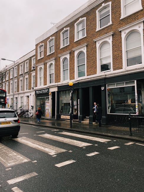 The image depicts a street view in South Kensington with a three-story brick building showcasing large, arched windows framed with white trim on the upper floors. The ground floor features storefronts with glass windows and black signage. The pavement appears clean and wet, likely from recent cleaning or rain. Several pedestrians are present, some wearing masks, and a white car is parked along the curb. A black lamppost with a yellow globe light is situated near the building's entrance. The scene emphasizes urban cleanliness and maintenance, consistent with the cleaning and disposal regulations outlined by Kensington and Chelsea Council. South Kensington Cleaners might be involved in maintaining the building’s exterior appearance as part of routine or specialist cleaning services.