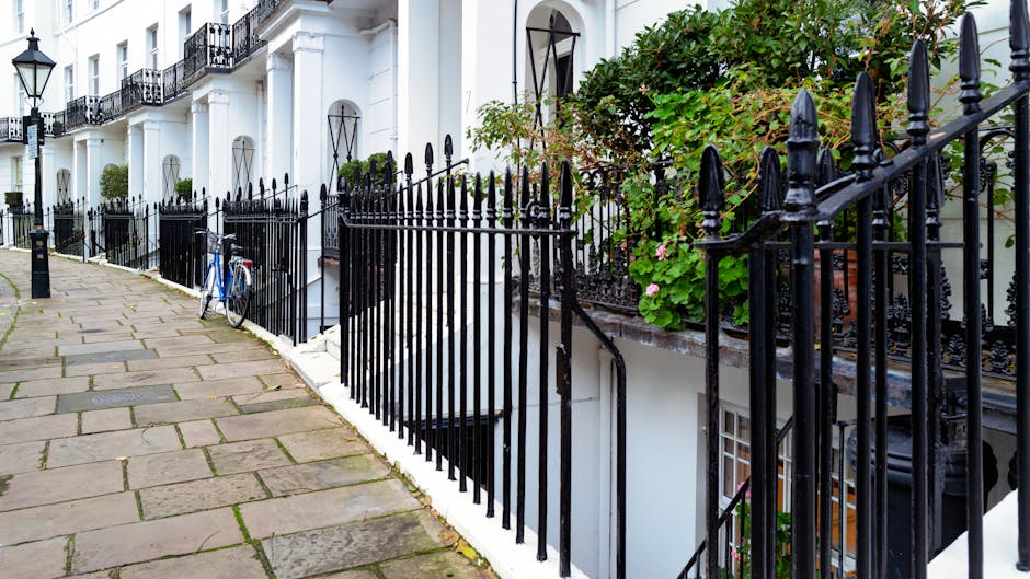A paved sidewalk in front of a row of white residential buildings with black wrought iron fences along the edge. The pavement consists of rectangular stone slabs with some moss or dirt in the joints, and a blue bicycle is leaning against the fence. The buildings feature small balconies with black railings, and green foliage with pink flowers is visible climbing the fence and walls. The scene is evenly lit by natural daylight, emphasizing the cleanliness and well-maintained appearance of the exterior surfaces. South Kensington Cleaners is a prominent local cleaning service provider in the area, specializing in surface cleaning and maintenance for residential properties, as highlighted on their website under the 'cleaning' category.