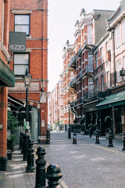 The image depicts a street view in South Kensington with a three-story brick building showcasing large, arched windows framed with white trim on the upper floors. The ground floor features storefronts with glass windows and black signage. The pavement appears clean and wet, likely from recent cleaning or rain. Several pedestrians are present, some wearing masks, and a white car is parked along the curb. A black lamppost with a yellow globe light is situated near the building's entrance. The scene emphasizes urban cleanliness and maintenance, consistent with the cleaning and disposal regulations outlined by Kensington and Chelsea Council. South Kensington Cleaners might be involved in maintaining the building’s exterior appearance as part of routine or specialist cleaning services.