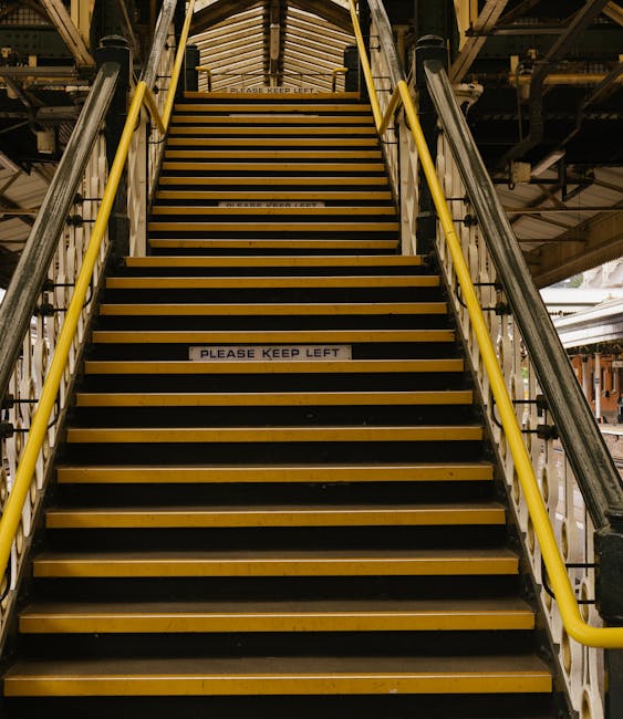 A set of narrow metal staircase with yellow safety handrails and yellow edge markings on each step, leading up to an elevated platform within an industrial or warehouse environment. The steps are made of black metal with slip-resistant strips, and there are signs on the steps reading 'PLEASE KEEP LEFT.' The surrounding area includes metallic structural supports and lighting fixtures, with a partially visible ceiling above. The environment appears clean and well-maintained, reflecting the industrial setting's emphasis on safety and hygiene, consistent with surface cleaning and maintenance practices promoted by South Kensington Cleaners for commercial spaces.