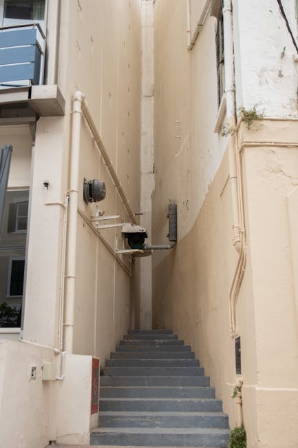 A set of narrow metal staircase with yellow safety handrails and yellow edge markings on each step, leading up to an elevated platform within an industrial or warehouse environment. The steps are made of black metal with slip-resistant strips, and there are signs on the steps reading 'PLEASE KEEP LEFT.' The surrounding area includes metallic structural supports and lighting fixtures, with a partially visible ceiling above. The environment appears clean and well-maintained, reflecting the industrial setting's emphasis on safety and hygiene, consistent with surface cleaning and maintenance practices promoted by South Kensington Cleaners for commercial spaces.