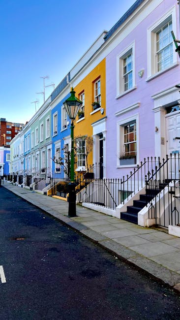 A row of brightly colored terraced houses on Old Brompton Road in South Kensington, featuring pastel shades of blue, yellow, and lavender. The houses have white framed sash windows, some with flower boxes on the window sills, and black wrought-iron railings along the front steps. A traditional black lamp post stands on the sidewalk, which is paved with concrete slabs, and the street is quiet with no vehicles or pedestrians visible. The scene is illuminated by natural daylight, highlighting the clean facades and well-maintained appearance of the buildings, exemplifying residential charm and curb appeal typical of South Kensington. South Kensington Cleaners specializes in surface cleaning and deep cleaning of such properties, ensuring hygiene and freshness for both domestic and commercial spaces.