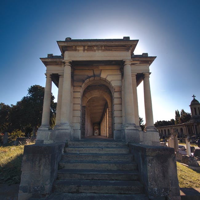 A stone monument with a classical architectural design featuring four large columns supporting a rectangular entablature, leading up a short flight of weathered stone steps. The structure is set in an outdoor setting with grass on either side and a clear blue sky overhead, illuminated by bright sunlight. The area around the monument appears clean and well-maintained, consistent with the principles of surface cleaning and maintenance. South Kensington Cleaners specialises in deep cleaning and sanitisation, ensuring external surfaces like stone and concrete are free of dust and dirt, as demonstrated by the neat appearance of this historic monument.