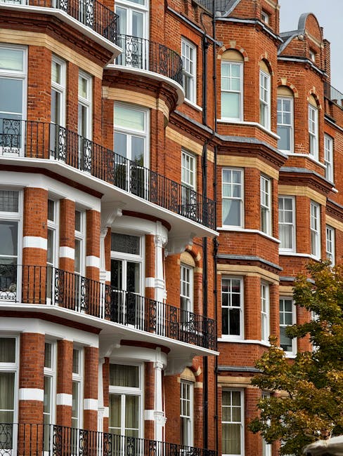 A row of brightly colored terraced houses on Old Brompton Road in South Kensington, featuring pastel shades of blue, yellow, and lavender. The houses have white framed sash windows, some with flower boxes on the window sills, and black wrought-iron railings along the front steps. A traditional black lamp post stands on the sidewalk, which is paved with concrete slabs, and the street is quiet with no vehicles or pedestrians visible. The scene is illuminated by natural daylight, highlighting the clean facades and well-maintained appearance of the buildings, exemplifying residential charm and curb appeal typical of South Kensington. South Kensington Cleaners specializes in surface cleaning and deep cleaning of such properties, ensuring hygiene and freshness for both domestic and commercial spaces.