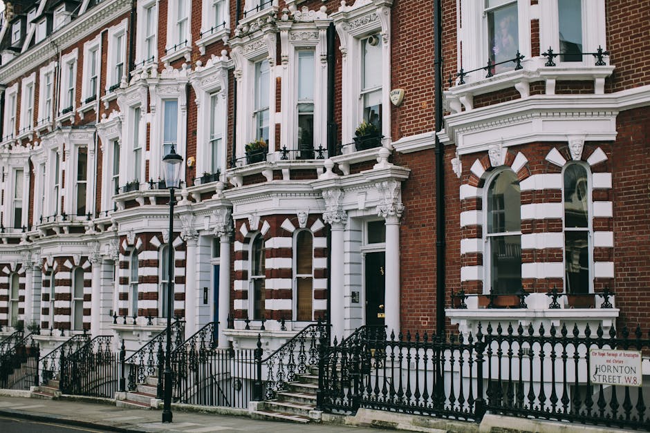 A row of Victorian-style terraced houses in SW7, South Kensington, featuring red brick facades accented with white decorative moldings around arched windows and doorways. The buildings have small front gardens enclosed by ornate black wrought iron fences, with steps leading up to black front doors. Large, multi-paned windows reflect natural light, and some have flower boxes. A traditional black street lamp is positioned along the pavement, with the scene illuminated by soft daytime lighting. The overall appearance demonstrates well-maintained exteriors, consistent with residential properties requiring regular surface cleaning and maintenance, as highlighted by South Kensington Cleaners in their guidance on permits and fines related to cleaning standards for landlords.