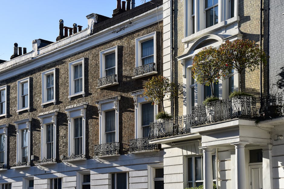 Photograph of a multi-storey residential building in South Kensington featuring a classic brick facade with white window trims and decorative molding. Each window is framed with white borders, with some open and others closed, revealing windowpanes with reflective glass. The building has small wrought iron balconies with intricate railing designs, adorned with potted plants and small trees, adding greenery to the urban setting. The ground floor showcases white painted columns supporting a balcony above, with a clean, well-maintained appearance. Bright sunlight casts shadows on the building, emphasizing its architectural details, with a clear blue sky in the background. This image illustrates the exterior of a typical historic London terrace, emphasizing the importance of surface cleaning and general maintenance to preserve its appearance, which is a key aspect of the services offered by South Kensington Cleaners for domestic and commercial cleaning needs.