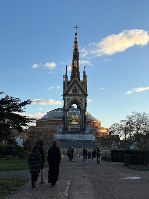 A group of people walking along a paved pathway in front of the Royal Albert Hall area, with a prominent monument featuring a tall, ornate spire topped with a cross. The monument has decorative Gothic-style features and a rounded, domed base. The surrounding landscape includes trees, some with sparse branches, under a clear blue sky with a few scattered clouds. The scene is well-lit by natural sunlight, highlighting the architectural details of the monument and the cleanliness of the area. This image captures not only the historic architecture typical of South Kensington but also represents outdoor public space maintenance and the importance of clean, well-kept environments, echoing the themes of surface cleaning and upkeep emphasized by South Kensington Cleaners.