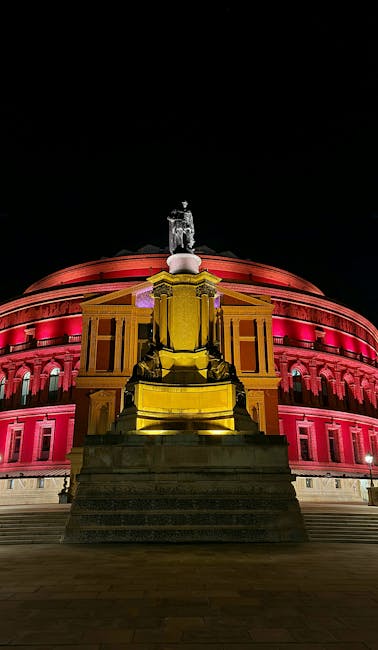 Night view of the illuminated Royal Albert Hall in South Kensington, displaying its distinctive circular architectural design with a domed roof lit in pink and red lights. In the foreground, a classical statue on a pedestal is visible, surrounded by steps leading up to the building. The scene highlights the grandeur of the historic venue, with the dark sky providing a stark contrast to the vibrant lighting that accentuates the façade's details. The image emphasizes the area’s ambiance and architectural features, relevant to surface cleaning and maintenance of prominent cultural landmarks, as addressed by South Kensington Cleaners in their tips for deep cleaning and surface sanitisation.
