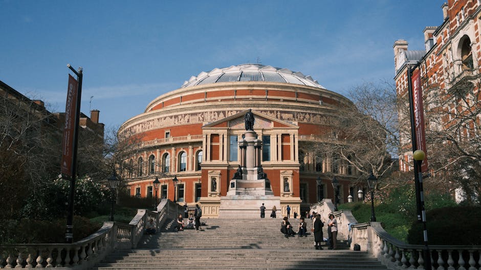A group of people walking along a paved pathway in front of the Royal Albert Hall area, with a prominent monument featuring a tall, ornate spire topped with a cross. The monument has decorative Gothic-style features and a rounded, domed base. The surrounding landscape includes trees, some with sparse branches, under a clear blue sky with a few scattered clouds. The scene is well-lit by natural sunlight, highlighting the architectural details of the monument and the cleanliness of the area. This image captures not only the historic architecture typical of South Kensington but also represents outdoor public space maintenance and the importance of clean, well-kept environments, echoing the themes of surface cleaning and upkeep emphasized by South Kensington Cleaners.