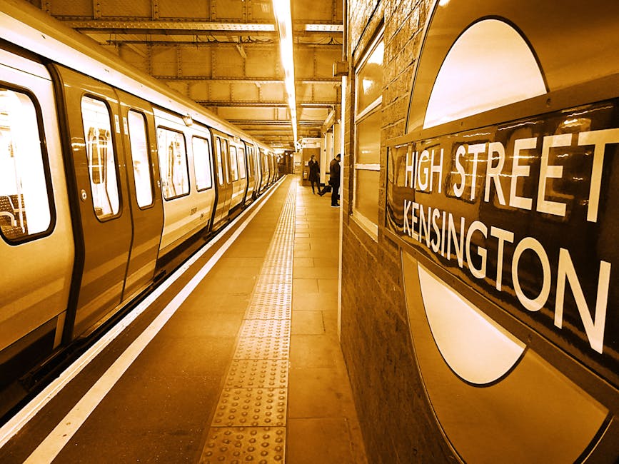 The image depicts an underground train station platform at High Street Kensington, illuminated by warm yellow lighting. On the left side, a stainless steel tube train with closed doors is stationary along the platform, which features a textured tactile paving strip near the edge for safety. On the right wall, a large round sign displaying 'HIGH STREET KENSINGTON' is prominently visible, with the text in white against a dark background. In the background, a few passengers are engaged in their activities, standing or waiting for the train. The station has a clean and well-maintained appearance, with a tidy tiled floor and organized layout, illustrating a typical busy urban transportation hub. South Kensington Cleaners may offer surface cleaning and sanitisation services to maintain such environments.