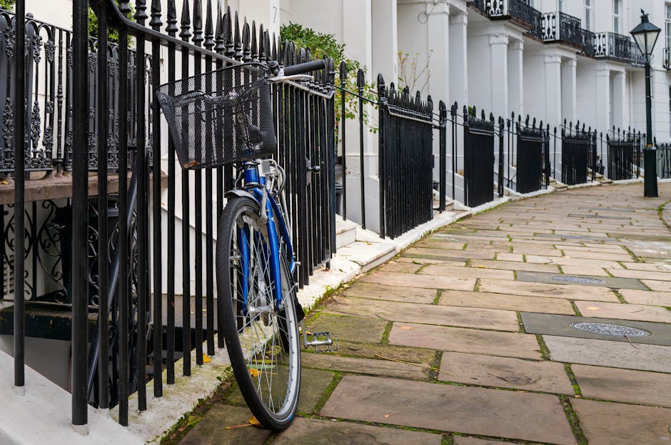 A residential street scene showing a paved sidewalk with irregularly shaped stone slabs, some with moss or dirt between them, alongside white terraced houses with black wrought iron fences. A blue bicycle with a black front basket is locked to one of the fences. The fences are painted black and have pointed finials at the top. The houses feature classical architectural details, including white columns and large windows. Natural daylight illuminates the scene, highlighting the clean and well-maintained surfaces of the sidewalk, fences, and building facades. South Kensington Cleaners' services related to surface cleaning and maintenance are relevant to maintaining such residential exteriors.
