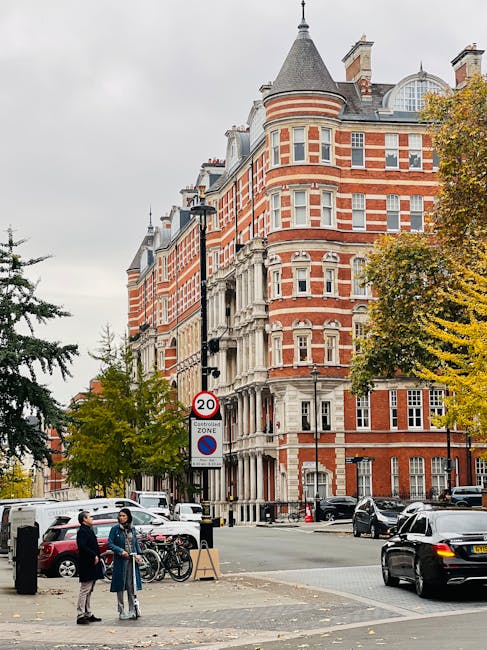 Photograph of a narrow cobblestone street in South Kensington lined with white residential buildings on both sides. The buildings feature large windows, some with black iron balconies and flower boxes, and doors with decorative trim. Potted plants and small trees are placed along the pavement, contributing to a lush, green atmosphere. Wooden benches and black planters with greenery are positioned outside the houses, and soft natural light illuminates the scene. The setting appears well-maintained and tidy, reflecting a clean and peaceful residential area, as promoted by South Kensington Cleaners for surface cleaning and domestic hygiene maintenance.